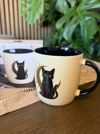 White ceramic mug with black interior and handle featuring a black cat sitting before a golden crescent moon with celestial detailing